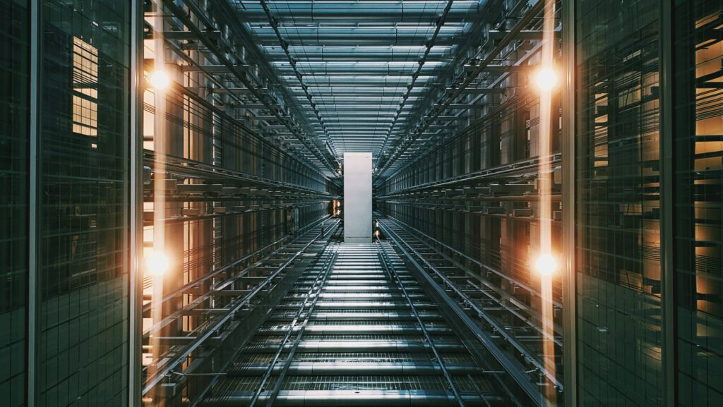 Blick nach oben durch einen hohen Gebäudeschacht mit symmetrischer Stahlkonstruktion, Glasdach und seitlichen Lichtquellen.
