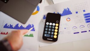 Smartphone mit geöffnetem Taschenrechner liegt auf ausgedruckten Diagrammen und Kennzahlen; eine Person zeigt mit einem Stift auf die dargestellten Auswertungen.