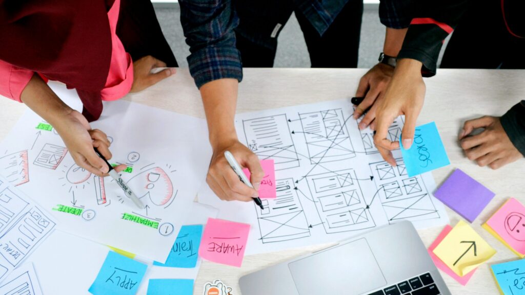 Mehrere Personen arbeiten gemeinsam an ausgedruckten Wireframes und Skizzen einer Benutzeroberfläche; auf dem Tisch liegen farbige Haftnotizen, Stifte und ein Laptop.