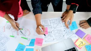 Mehrere Personen arbeiten gemeinsam an ausgedruckten Wireframes und Skizzen einer Benutzeroberfläche; auf dem Tisch liegen farbige Haftnotizen, Stifte und ein Laptop.